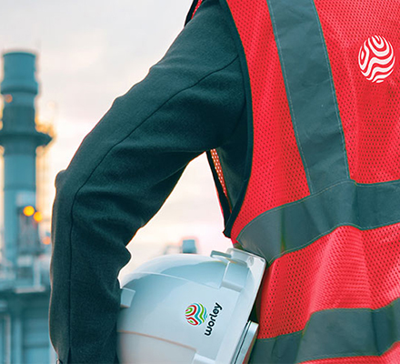 Person wearing PPE holding a Optravation Energysafety helmet under his arm in front of a refinery.